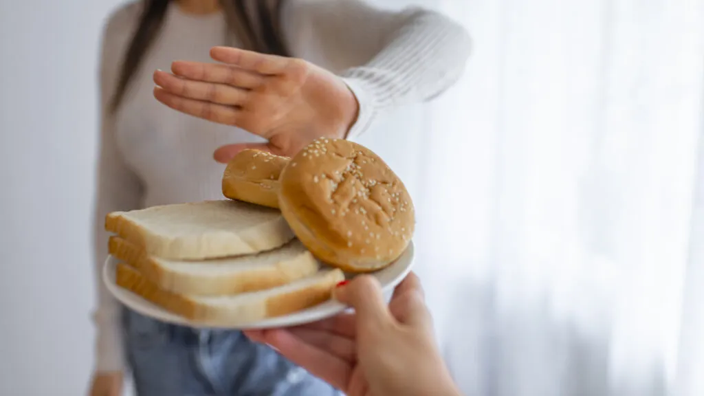 Kobieta podaje talerz z dużą ilością kromek chleba, ale druga kobieta odmawia ich przyjęcia - w nawiązaniu do węglowodanów i chudnięcia