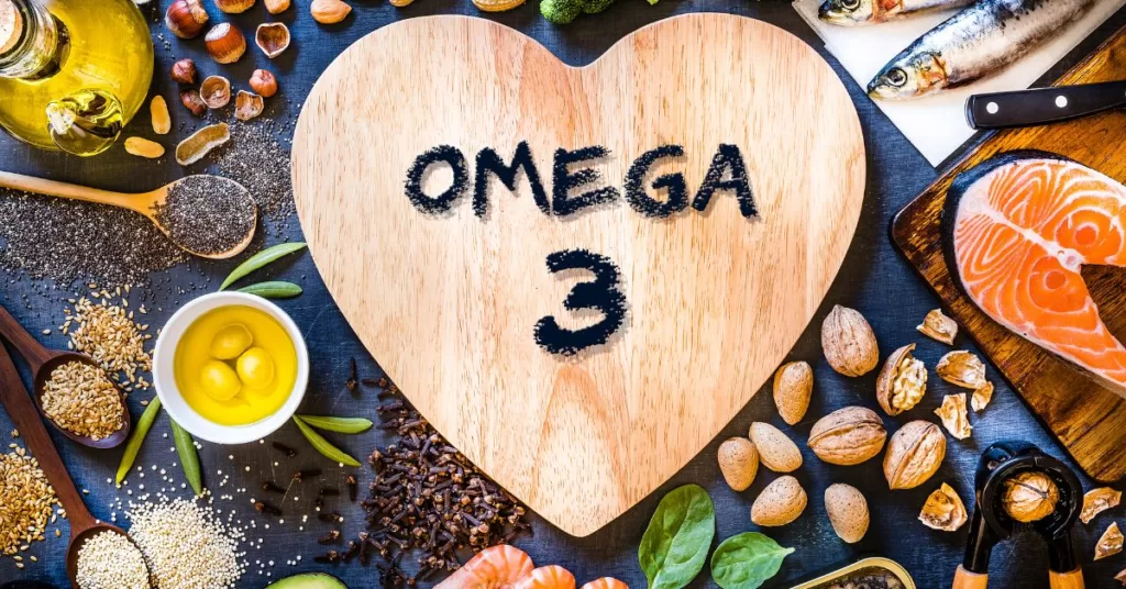 Deska w kształcie serca na którym widnieje napis "Omega 3", a obok znajdują się ryby morskie, orzechy i inne źródła NNKT