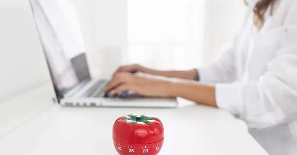 Kobieta pracuje za pomocą laptopa, a obok niej leży minutnik w kształcie pomidora